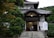 A traditional Japanese building with intricate wooden architecture and a tiled roof, surrounded by lush green trees. The entrance features a small flight of stairs leading to wooden doors and signposts with Japanese inscriptions.