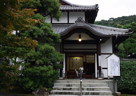 A traditional Japanese building with intricate wooden architecture and a tiled roof, surrounded by lush green trees. The entrance features a small flight of stairs leading to wooden doors and signposts with Japanese inscriptions.
