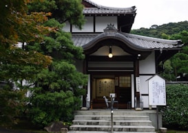 A traditional Japanese building with intricate wooden architecture and a tiled roof, surrounded by lush green trees. The entrance features a small flight of stairs leading to wooden doors and signposts with Japanese inscriptions.