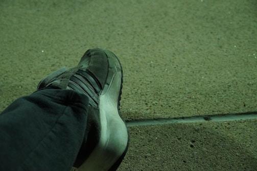A single sneaker resting against a rough-textured concrete surface with a slight green tint. The shoe is dark with a prominent rubber sole and features several stripes along the side.