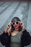 A woman with stylish sunglasses and striking lipstick poses confidently, holding the white frames with her fingers. Her top is an olive green, one-shouldered design, and she wears a black jacket with puffy sleeves. Her silver wristwatch complements her look, while her nails are painted red. Her hair is medium-length with highlights, and she stands against a soft, blurred background.