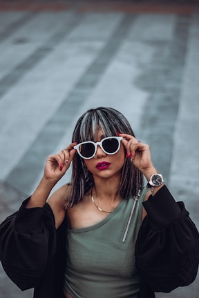 A woman with stylish sunglasses and striking lipstick poses confidently, holding the white frames with her fingers. Her top is an olive green, one-shouldered design, and she wears a black jacket with puffy sleeves. Her silver wristwatch complements her look, while her nails are painted red. Her hair is medium-length with highlights, and she stands against a soft, blurred background.