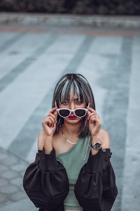 A person with highlighted hair holds a pair of white-framed sunglasses in front of their eyes. They are wearing a one-shoulder green top paired with an off-the-shoulder black jacket and a wristwatch on their left wrist. The background features a patterned pavement in muted tones of gray and blue.