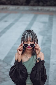 A person with highlighted hair holds a pair of white-framed sunglasses in front of their eyes. They are wearing a one-shoulder green top paired with an off-the-shoulder black jacket and a wristwatch on their left wrist. The background features a patterned pavement in muted tones of gray and blue.