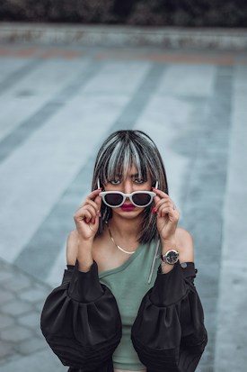 A person with highlighted hair holds a pair of white-framed sunglasses in front of their eyes. They are wearing a one-shoulder green top paired with an off-the-shoulder black jacket and a wristwatch on their left wrist. The background features a patterned pavement in muted tones of gray and blue.
