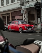 A vintage red car is parked on the street in front of a shop labeled Ice Cream & Coffee. The car stands out with its classic design and polished bodywork, and the setting includes several parked scooters and bicycles. The backdrop is an older building with a few people visible near the entrance.