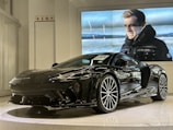 A sleek black sports car is displayed in a showroom with a modern and polished design. Behind the car, a large video screen shows a person in a black jacket standing in a cold outdoor setting, with snowy landscapes in the background.