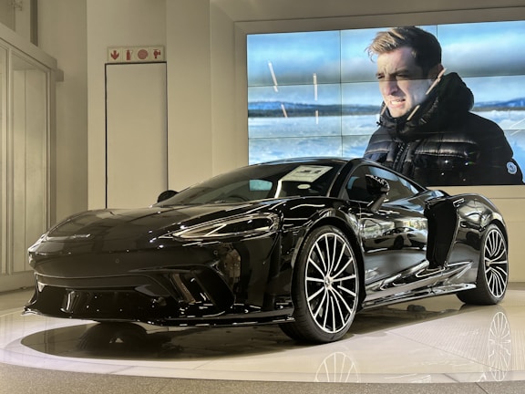 A sleek black sports car is displayed in a showroom with a modern and polished design. Behind the car, a large video screen shows a person in a black jacket standing in a cold outdoor setting, with snowy landscapes in the background.
