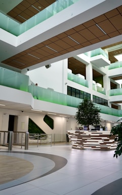 A modern interior with multiple levels, featuring sleek white walls and wood paneling on the ceilings. Glass balconies with green accents overlook the open space, and a central area contains a reception desk with a contemporary design. The area is adorned with indoor plants, contributing to a fresh and inviting atmosphere.