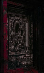 An intricately designed religious icon featuring figures with darkened faces, surrounded by ornate patterns and textures. Adjacent is a tall, decorative candle holder. The overall style appears to be historical and baroque, set against a backdrop with rich, dark colors.