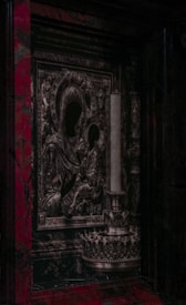 An intricately designed religious icon featuring figures with darkened faces, surrounded by ornate patterns and textures. Adjacent is a tall, decorative candle holder. The overall style appears to be historical and baroque, set against a backdrop with rich, dark colors.