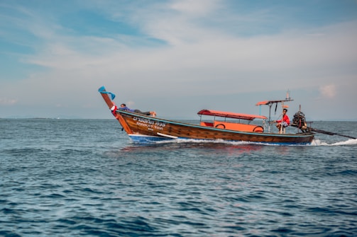 A small wooden boat with an orange canopy sails across a calm blue ocean. The boat has lettering on its side and a person steering at the rear. The sky is partly cloudy with a light blue hue.