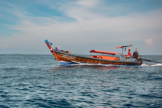 A small wooden boat with an orange canopy sails across a calm blue ocean. The boat has lettering on its side and a person steering at the rear. The sky is partly cloudy with a light blue hue.