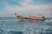 A small wooden boat with an orange canopy sails across a calm blue ocean. The boat has lettering on its side and a person steering at the rear. The sky is partly cloudy with a light blue hue.