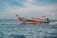 A small wooden boat with an orange canopy sails across a calm blue ocean. The boat has lettering on its side and a person steering at the rear. The sky is partly cloudy with a light blue hue.