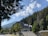 A picturesque scene featuring a large house with a unique roof design, nestled among a dense forest of pine trees. The backdrop includes a scenic view of mountains under a clear sky dotted with fluffy white clouds. A wooden fence runs along the bottom of the image, and a paved road is visible on the left side.