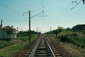 Railway tracks intersect and diverge in a rural setting, surrounded by green grass and vegetation. An old building with a tiled roof stands to the left, while electrical poles and power lines extend into the distance. The scene is peaceful with a slightly overcast sky and distant hills.