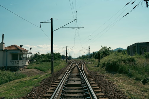 Railway tracks intersect and diverge in a rural setting, surrounded by green grass and vegetation. An old building with a tiled roof stands to the left, while electrical poles and power lines extend into the distance. The scene is peaceful with a slightly overcast sky and distant hills.