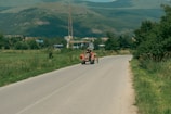 A rural road flanked by fields and trees leads toward a distant town with a backdrop of green mountains. A tractor with two people on board travels along the road. In the background, there are structures like a gas station and communication tower.