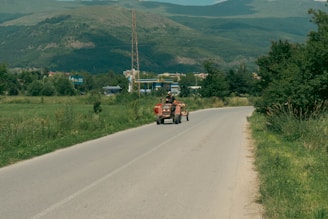 A rural road flanked by fields and trees leads toward a distant town with a backdrop of green mountains. A tractor with two people on board travels along the road. In the background, there are structures like a gas station and communication tower.