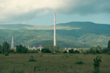 A large industrial chimney rises against a backdrop of expansive green hills and forests under a soft, cloudy sky. The foreground features an open grassy field with scattered shrubs, while the industrial complex is partially visible, surrounded by trees.