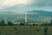 A large industrial chimney rises against a backdrop of expansive green hills and forests under a soft, cloudy sky. The foreground features an open grassy field with scattered shrubs, while the industrial complex is partially visible, surrounded by trees.