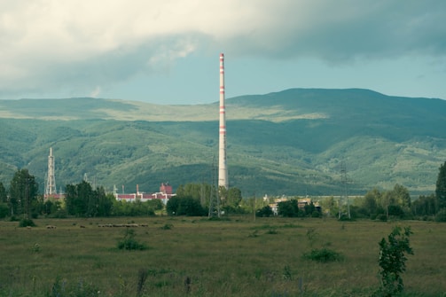 A large industrial chimney rises against a backdrop of expansive green hills and forests under a soft, cloudy sky. The foreground features an open grassy field with scattered shrubs, while the industrial complex is partially visible, surrounded by trees.