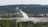 A geyser is erupting in the middle of a wooded landscape, surrounded by dense forest. The scene includes several buildings in the distance, indicating a tourist or visitor center area. A road or pathway encircles the geyser, with what appears to be people gathered to observe the natural spectacle.