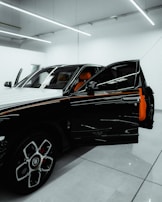 A luxury car is parked in a modern showroom with bright overhead lighting. The driver’s door is open, revealing a vibrant orange interior. The car's exterior features a sleek, black-and-white design with distinctive rims.