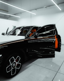 A luxury car is parked in a modern showroom with bright overhead lighting. The driver’s door is open, revealing a vibrant orange interior. The car's exterior features a sleek, black-and-white design with distinctive rims.
