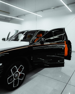 A luxury car is parked in a modern showroom with bright overhead lighting. The driver’s door is open, revealing a vibrant orange interior. The car's exterior features a sleek, black-and-white design with distinctive rims.
