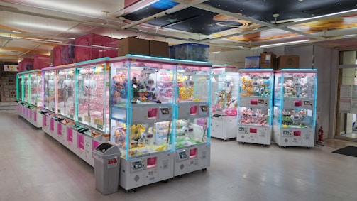 A bright and colorful arcade filled with numerous claw machines lined up in rows. Each machine is filled with plush toys and prizes, illuminated with neon blue and pink lights. The floor is clean and shiny, and there's a visible trash bin near the machines. The ceiling has decorative panels with various patterns.