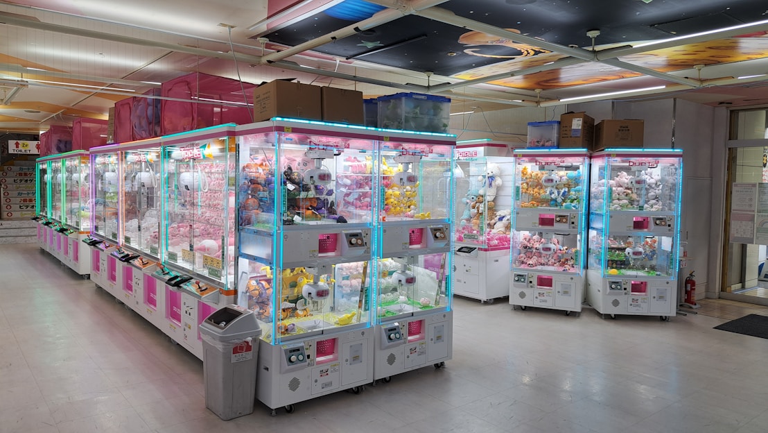 A colorful arcade scene featuring various claw machines including mini, medium, and giant sizes with plush toys inside.
