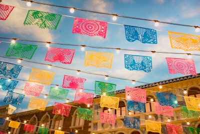 Colorful papel picado banners are strung across the scene, illuminated by hanging string lights against a background of blue sky and a building with classical architecture.