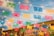 Colorful papel picado banners are strung across the scene, illuminated by hanging string lights against a background of blue sky and a building with classical architecture.