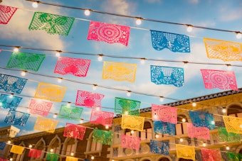 Colorful papel picado banners are strung across the scene, illuminated by hanging string lights against a background of blue sky and a building with classical architecture.