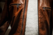A close-up view of a brown leather jacket featuring two zippered pockets on the chest, worn over a textured white knit sweater.