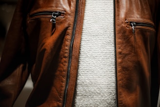 A close-up view of a brown leather jacket featuring two zippered pockets on the chest, worn over a textured white knit sweater.