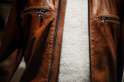 A close-up view of a brown leather jacket featuring two zippered pockets on the chest, worn over a textured white knit sweater.