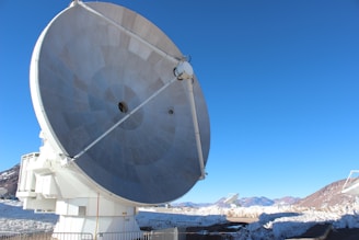 A large, metallic radio telescope dish is positioned prominently in the foreground against a clear blue sky. Snow-covered mountains and dry terrain are visible in the background, and additional satellite dishes can be seen in the distance. The scene is enclosed with metal barriers around the base of the dish.