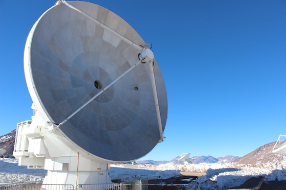 A large, metallic radio telescope dish is positioned prominently in the foreground against a clear blue sky. Snow-covered mountains and dry terrain are visible in the background, and additional satellite dishes can be seen in the distance. The scene is enclosed with metal barriers around the base of the dish.