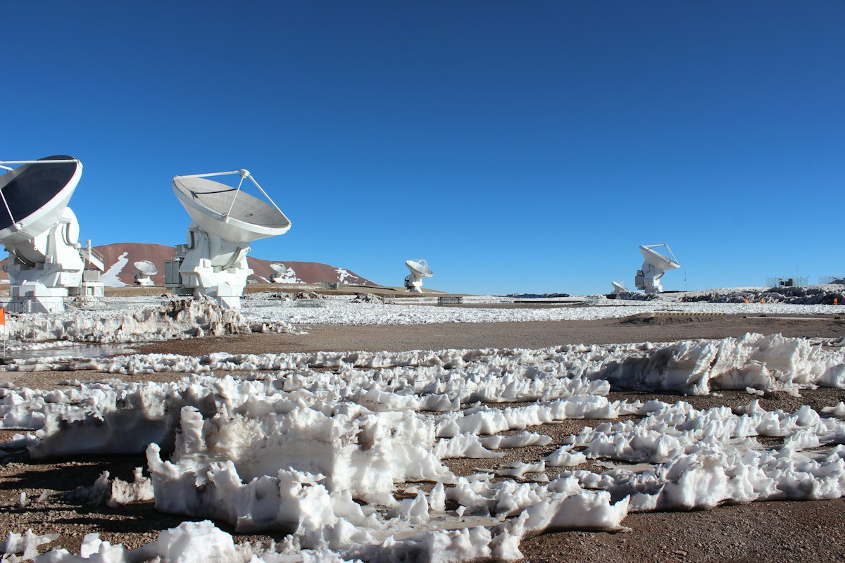 Four white radio telescope dishes on a high, dry plain under an intense blue sky: two in the foreground on their transporter mounts, two more distant on the horizon. The ground is brown and bare, with melting white blocks of snow forming a rough band across the front, and a reddish hill in the background