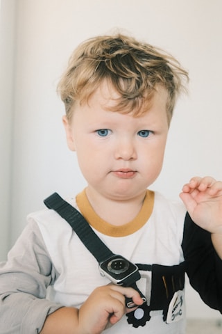 A young child with light hair and blue eyes wearing a long-sleeved shirt. The shirt has a yellow neckline and what appears to be a toy truck design. The child is holding a black strap with a small object attached to it, possibly a toy watch.