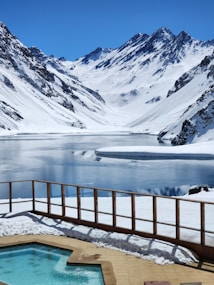 A serene mountain landscape with snow-covered peaks and a tranquil, partially frozen lake. In the foreground, there is a hot tub with clear blue water, surrounded by a wooden railing and snow. The sky is a deep blue, enhancing the crispness of the snow and the peaceful atmosphere.