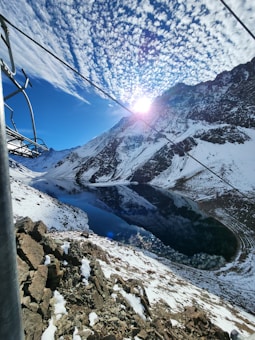 The image features a breathtaking mountainous landscape with a large, calm lake reflecting the rugged, snow-capped mountains surrounding it. The sky is filled with a pattern of scattered clouds, and the bright sun peeks through, casting a brilliant glow over the scene. There is a metal structure and cables visible on the left side of the image, possibly part of a ski lift or cable car system.