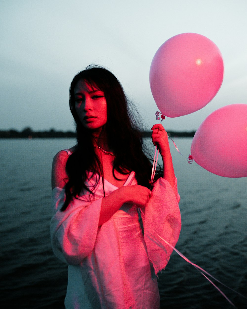 a woman holding three pink balloons in front of a body of water