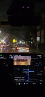 Nighttime inside a car with a visible dashboard displaying a GPS navigation system. The outside view through the windshield shows a street illuminated by traffic lights and car headlights. Road signs are visible, including a speed limit sign indicating 35 mph.