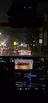 Nighttime inside a car with a visible dashboard displaying a GPS navigation system. The outside view through the windshield shows a street illuminated by traffic lights and car headlights. Road signs are visible, including a speed limit sign indicating 35 mph.