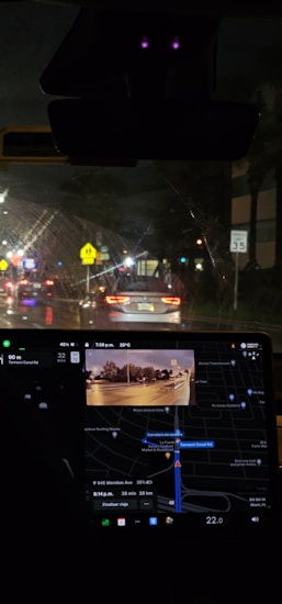 Nighttime inside a car with a visible dashboard displaying a GPS navigation system. The outside view through the windshield shows a street illuminated by traffic lights and car headlights. Road signs are visible, including a speed limit sign indicating 35 mph.
