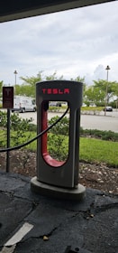A Tesla charging station situated outdoors, with the brand name illuminated in red. Vegetation and a parking lot with several vehicles are visible in the background under a cloudy sky.